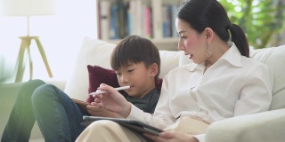 多莉展示了一个富有的亚洲妇女和她的儿子一起在客厅使用数码平板电脑的照片，开心的微笑。