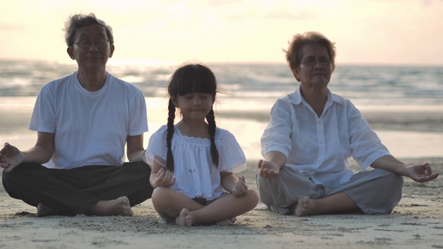 日落时，在沙滩上做瑜伽的爷爷奶奶和孙女的剪影。家庭，生活方式，人，人寿保险，多代人，老人，假期，关系，孩子，假期，退休，健康护理，领导理念。