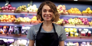 年轻漂亮的女售货员穿着黑色围裙站在超市货架上的水果在背景，看着相机和微笑。商贸以人为本的理念