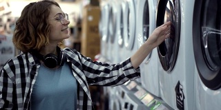 一个穿着格子衬衫的年轻女人正在家电商店里挑选洗衣机。打开门往里看。侧视图