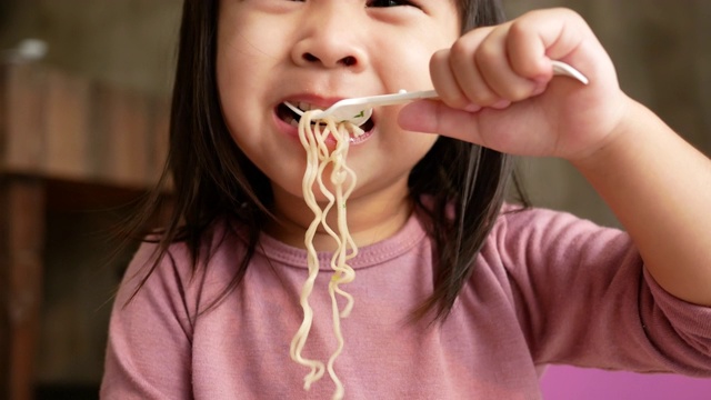 亚洲小女孩有乐趣吃面条在塑料杯在家里。垃圾食品的概念。