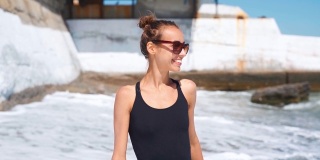慢动作苗条性感健美微笑的年轻女子在黑色泳衣走在海边与波浪
