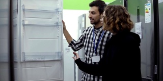 在一家消费电子商店购买前，一名年轻女子站在打开冰箱的门，与一名男性顾问讨论冰箱的设计和质量。与商店顾问讨论特点