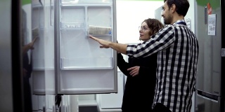 年轻的夫妇在消费电子商店购买前检查开门式冰箱，设计和质量。讨论特点及设计