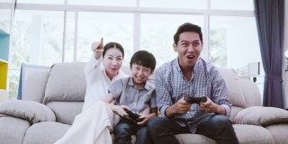 闲暇的日子和爱好亚洲家庭，爸爸妈妈和儿子坐在沙发上看电视玩电子游戏快乐的乐趣在客厅里的房子