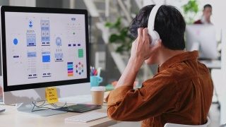 在现代创意办公室，年轻的亚洲程序员男子戴着耳机在电脑桌面上编写移动应用程序，忙碌的亚洲ux, ui设计开发团队，放松的亚洲男性听着音乐工作视频素材模板下载