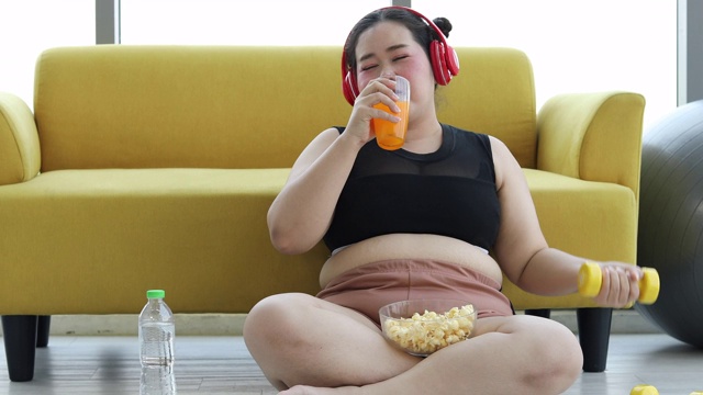 大块头女人听着音乐和亚洲女孩喜欢在家里的沙发上吃东西，运动减肥为肥胖女人的概念