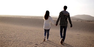 这是罕见的一对情侣在阳光下和沙漠里的蓝天下牵手浪漫地散步。两个年轻的恋人，穿着休闲的衣服，手牵着手走在沙漠的沙子上。镜头耀斑