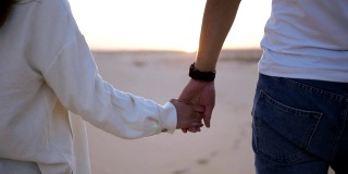 镜头中，这对牵手的情侣在阳光下和沙漠中的蓝天下浪漫地散步。两个年轻的恋人穿着休闲的衣服漫步在沙漠的沙滩上。景观上的背景