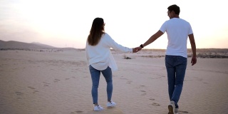 背后的镜头牵手的夫妇在阳光下和沙漠的蓝天下走在浪漫的关系。两个年轻的恋人穿着休闲的衣服漫步在沙漠的沙滩上。景观背景。慢动作