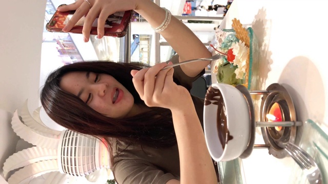 亚洲少女使用智能手机进行网络摄像头，在咖啡店吃冰淇淋，垂直格式的视频智能手机