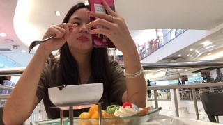 亚洲少女一边在咖啡店吃冰淇淋，一边用智能手机进行网络摄像头，身体状况良好，竖起大拇指视频素材模板下载