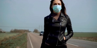 近女子在保护面罩和黑色夹克步行过马路停车与车轮上的行李箱和汽车经过