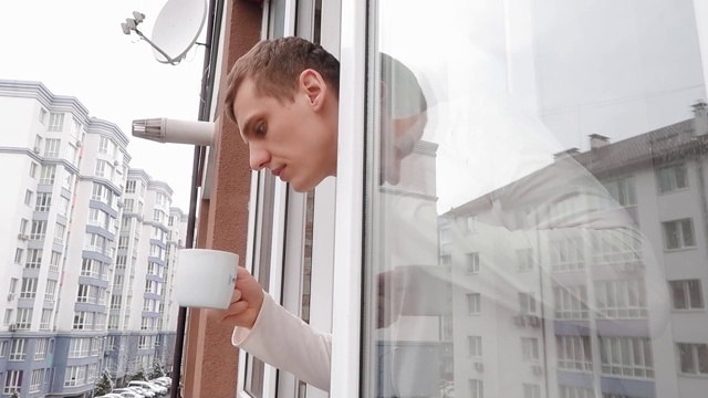 年轻人望着窗外的街道，用一只白色的杯子喝了一口咖啡或茶。隔离期间在家隔离。