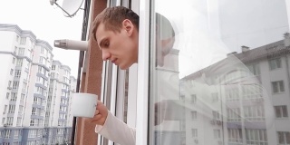 年轻人望着窗外的街道，用一只白色的杯子喝了一口咖啡或茶。隔离期间在家隔离。