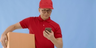亚洲快乐快递员戴着帽子和红色衬衫使用智能手机，带着纸包裹盒子孤立在蓝色背景和微笑的脸。邮政投递服务的概念。