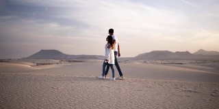在沙漠中，阳光下，蓝天下，一对情侣浪漫地走在一起的慢镜头。两个年轻的恋人穿着休闲的衣服漫步在沙漠的沙滩上。完美模糊的背景山的场景