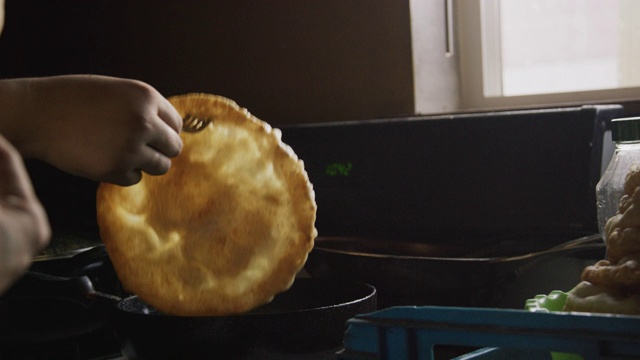 一个印第安人(纳瓦霍人)的女人的手形成一个生的玉米粉圆饼(炸面包)，而另一个女人取出一个熟的玉米粉圆饼，并把它放在一个完成的堆栈上，然后把一个生玉米粉圆饼放到室内炉子上的油