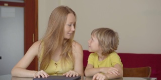 一位年轻妇女和她的小儿子在隔离期间坐在家里，在互联网学校使用平板电脑学习。冠状病毒的概念