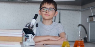 一个男生坐在厨房里的显微镜和课本旁边，看着摄像机微笑着