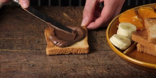 一个女人的手用一把铁刀在一块烤面包上涂上巧克力酱，然后在上面放上香蕉片