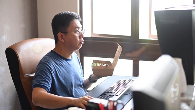 一个亚洲华人中年男子正在吃午餐，在他的家庭办公室工作的时候吃他的午餐