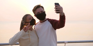 在美丽的日出时分，幸福的年轻夫妇戴着防护面具在海边用智能手机自拍