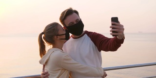 幸福的年轻夫妇戴着防护面具，在美丽的日出时用智能手机在海边自拍，慢镜头
