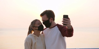 快乐的年轻夫妇戴着防护面具在美丽的日出期间在海边自拍和接吻