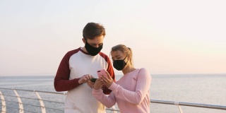 幸福的年轻夫妇戴着防护面具在美丽的日出期间在海边使用手机