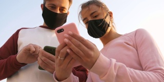 快乐的年轻夫妇戴着防护面具在海边用手机户外看美丽的日出，低角度的视野