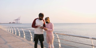 快乐的年轻夫妇戴着防护面具在美丽的日出时在海边使用手机，慢镜头