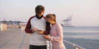 在美丽的日出期间，快乐的年轻夫妇戴着防护面具在海边使用手机