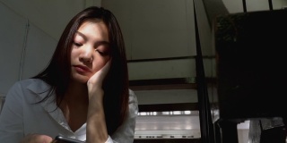 沮丧的年轻美丽的亚洲女人独自坐在楼梯上，用智能手机上网。有压力的孤独少女盯着手机带着负面情绪，然后闭上眼睛。