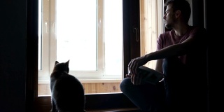 一个有魅力的男人和一只可爱的猫坐在房子的窗台上看书。自我隔离、隔离、大流行概念。