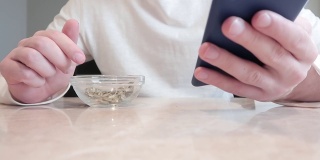 一名年轻人正在吃透明碗里的南瓜子，手里拿着智能手机