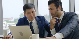 两名商人正坐在一个会议上，看着一台笔记本电脑讨论如何在公司现代化的办公室里帮助规划商业成功