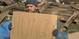 一个无家可归的人在废墟附近拿着饥饿的指示牌，在疫情期间帮助贫穷和饥饿的人们