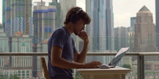 一个年轻的自由职业者在阳台上用笔记本电脑工作，背景是一个摩天大楼林立的城市中心。自由职业者it专家解决客户的问题