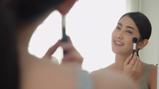 美女化妆刷用粉状化妆品涂抹面部肌肤。快乐自信的亚洲美丽的年轻女子化妆用粉敷在脸上在镜子在家里早上。