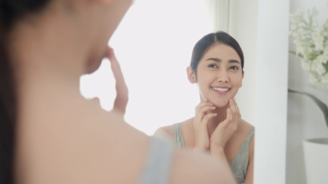 快乐自信的亚洲美丽的年轻女子看着脸微笑和脸在卧室镜子在每天例行在家里。