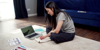 从侧面看亚洲女学生坐在家里的地板上，在线学习，观看与老师的视频会议，并在笔记本上做笔记