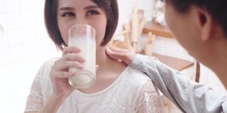 亚洲新夫妇家庭住在房子里。孕妇坐在沙发上捧着牛奶杯，丈夫给女孩牛奶然后女孩在家喝牛奶，开心地笑着看着他。婴儿家庭怀孕概念