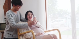 亚洲新夫妇家庭住在房子里。孕妇坐在沙发上捧着牛奶杯，丈夫给女孩牛奶然后女孩在家喝牛奶，开心地笑着看着他。婴儿家庭怀孕概念
