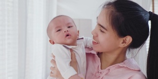 亚洲美丽的母亲在家里的卧室里温柔地拥抱着小女婴，然后亲吻年幼的孩子。在家庭关系中，温柔的触摸，关爱，保护妈妈和孩子。