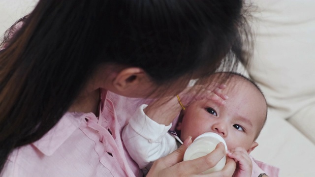 美丽的亚洲母亲在家里用奶瓶喂她的婴儿。婴儿躺在她的腿上。从俯视图透过母亲的肩膀，而孩子看着她。