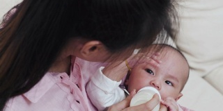 美丽的亚洲母亲在家里用奶瓶喂她的婴儿。婴儿躺在她的腿上。从俯视图透过母亲的肩膀，而孩子看着她。