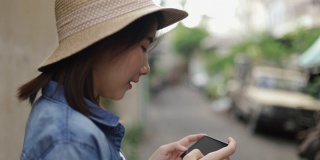 一名亚洲女子戴着复古软呢帽，在泰国曼谷的一条小街上用智能手机拍照，享受东南亚暑假旅游假期。