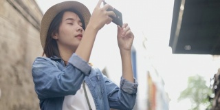 泰国曼谷的一条小街上，漂亮的潮女人戴着复古软呢帽，用智能手机拍照，正在享受东南亚的暑假旅游假期。