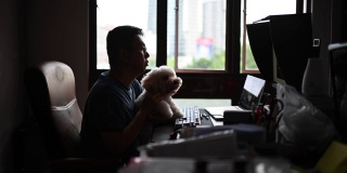 一位亚洲华人中年男子在网上与朋友、家人和商业伙伴聊天、聊天，一边在家里办公室的电脑屏幕上看着摄像头聊天，一边带着他的玩具狮子狗